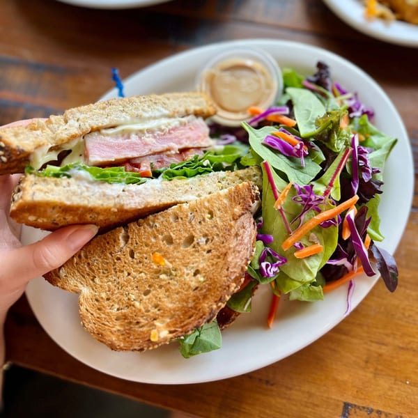 A hand holds a Lazy Daisy breakfast sandwich with ham, lettuce, and tomato, next to a side salad.