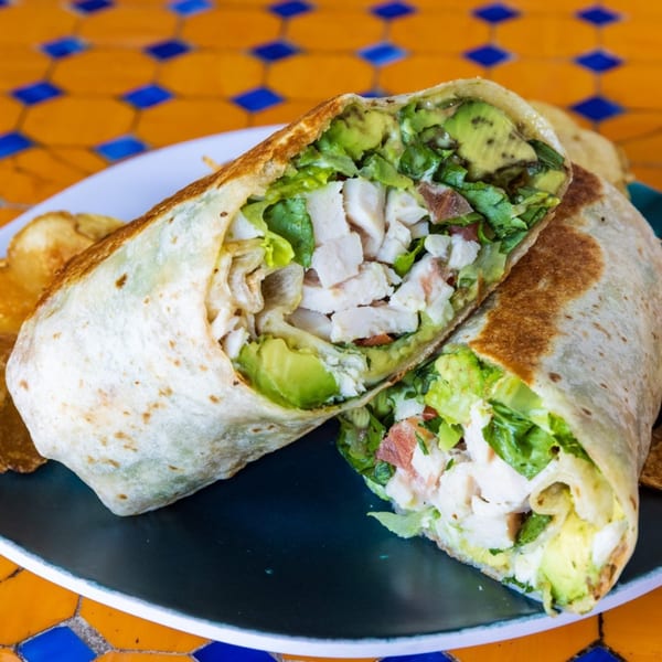 A chicken avocado wrap, cut in half, revealing chicken, avocado, lettuce, and bacon, on a plate.