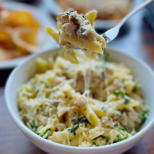 Fork lifting creamy penne pasta with chicken, cheese, and herbs from a white bowl.