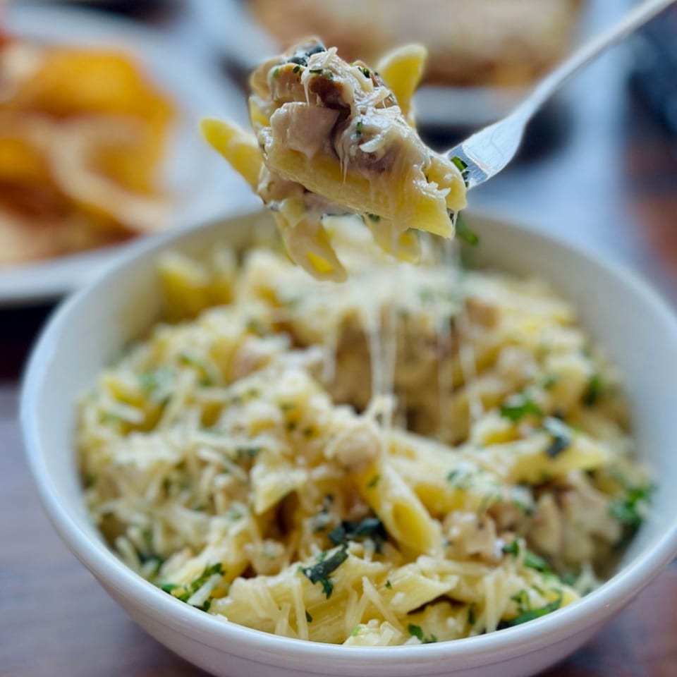 Fork lifting creamy penne pasta with chicken, cheese, and herbs from a white bowl.