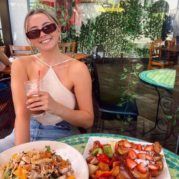 A smiling woman with sunglasses enjoys breakfast at Lazy Daisy, featuring French toast and a salad.