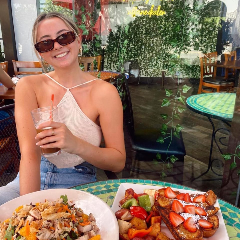 A smiling woman with sunglasses enjoys breakfast at Lazy Daisy, featuring French toast and a salad.