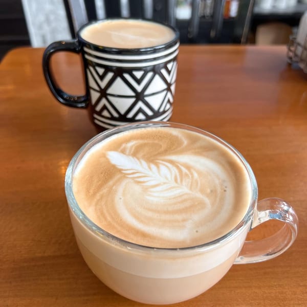 Two Lazy Daisy lattes on a wooden table, one with latte art in a clear mug.