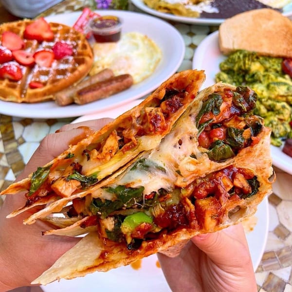 Hands holding a breakfast quesadilla filled with chicken, spinach, and cheese, with other breakfast plates in the background.