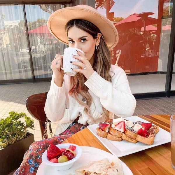 A woman in a hat sips coffee at Lazy Daisy, with French toast and fruit on the table.