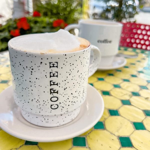 Two coffee cups, one with frothy foam, on a yellow and green tiled outdoor table.