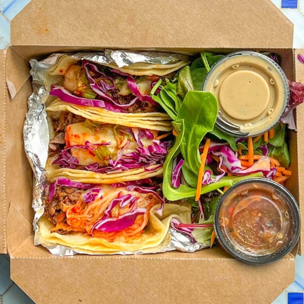Three breakfast tacos with purple cabbage, kimchi, and meat, alongside a side salad and two sauces.