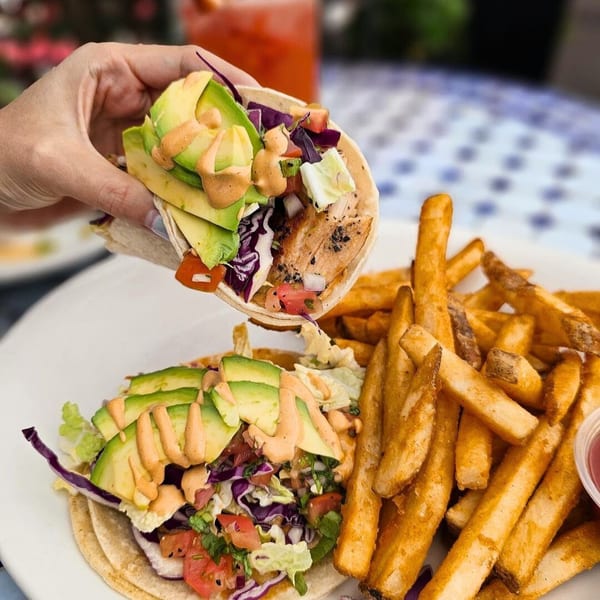 Hand holding a fish taco with avocado and sauce over another taco and a side of fries.