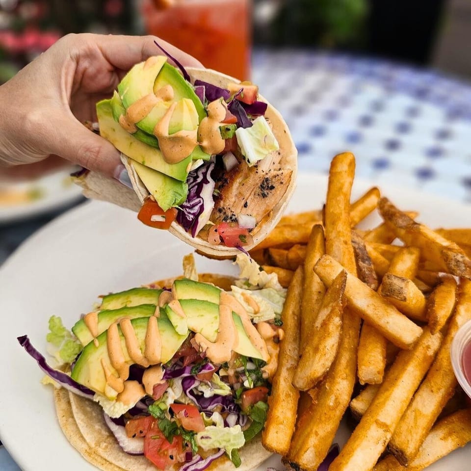 Hand holding a fish taco with avocado and sauce over another taco and a side of fries.