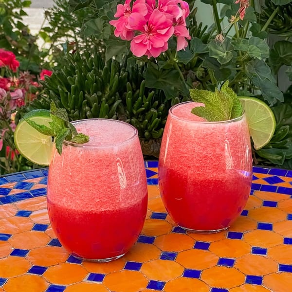 Two watermelon drinks with lime and mint garnishes on a colorful tiled table with pink flowers.