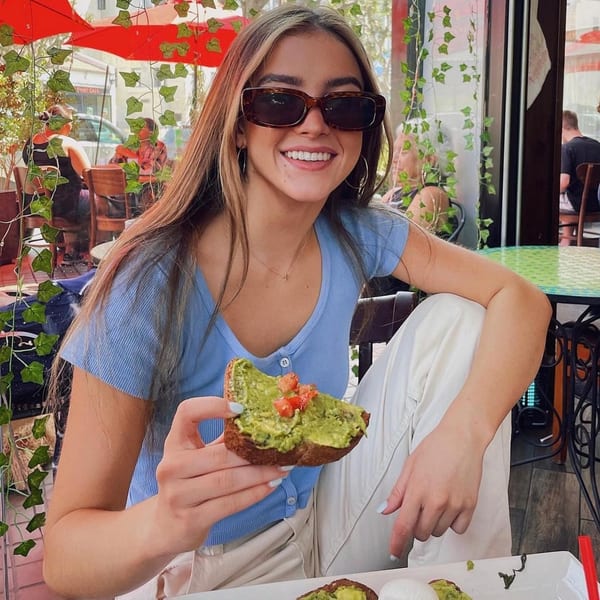 Smiling woman in sunglasses holding avocado toast with tomatoes at Lazy Daisy restaurant.