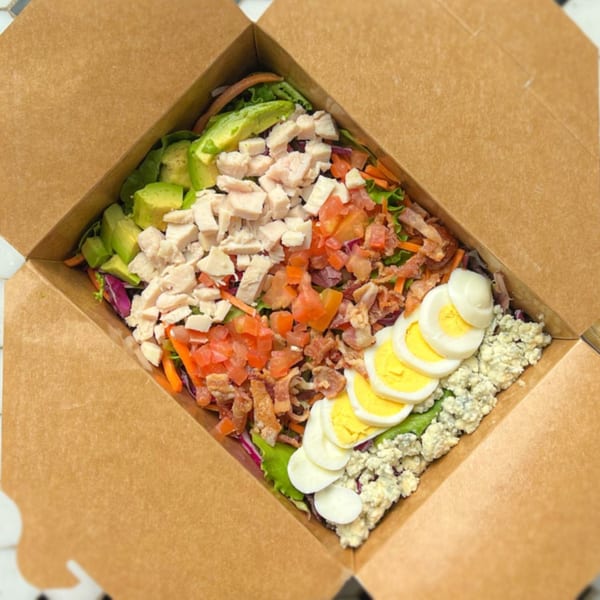 A Lazy Daisy Cobb salad in a brown takeout box, with avocado, chicken, tomatoes, bacon, eggs, and blue cheese.