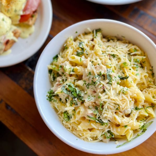 A white bowl of creamy penne pasta with shredded cheese and fresh herbs, next to eggs benedict.