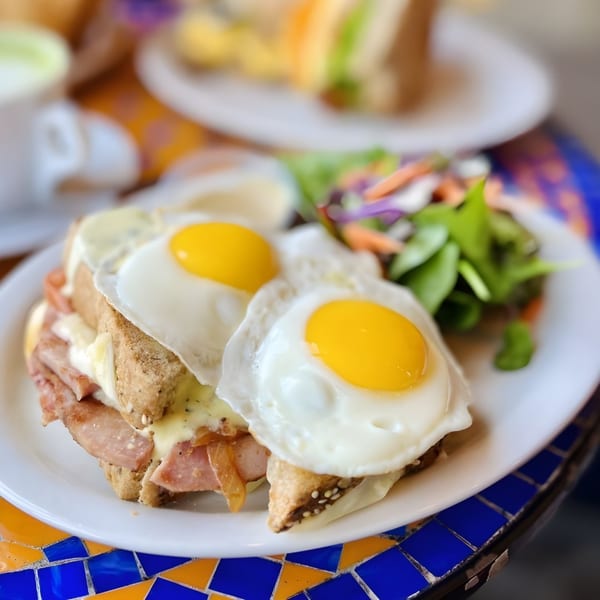 Lazy Daisy's Croque Madame with two fried eggs and a side salad on a colorful mosaic table.